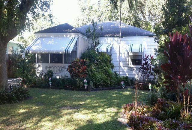 A white house with striped awnings sits behind a lush green lawn and tropical landscaping — 44 SW 5th Street, Dania Beach FL 33004, DANIA BEACH, FL