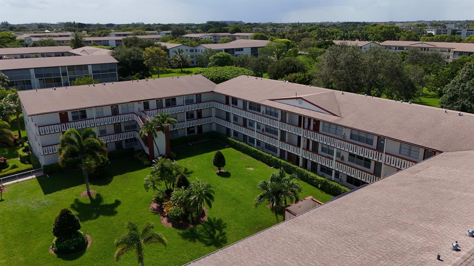 An aerial view of a multi-story apartment complex surrounding a central grassy courtyard with palm trees — 328 Brighton # H, Boca Raton FL 33434, BOCA RATON, FL