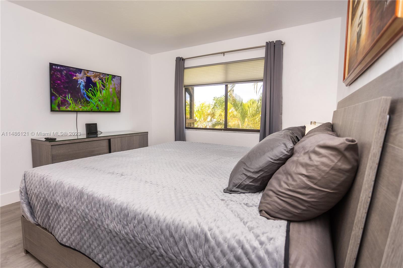 A bedroom features a gray bed, a wall-mounted television, and a window with gray curtains — 500 N Francisco St # 234, Clewiston FL 33440, CLEWISTON, FL