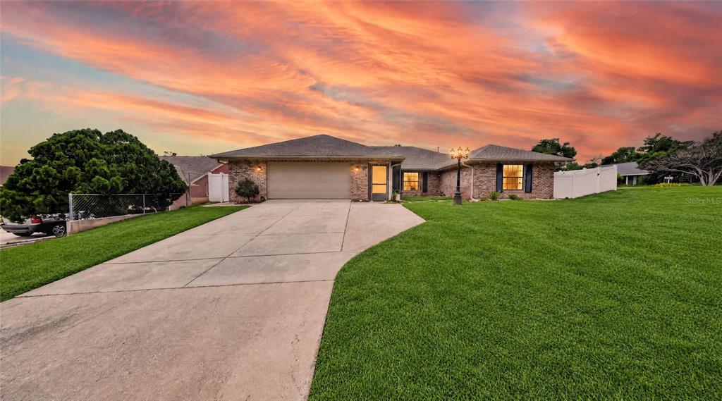 A single-story brick house with a concrete driveway and a large green lawn at sunset — 937 UTICA STREET, DELTONA, FL