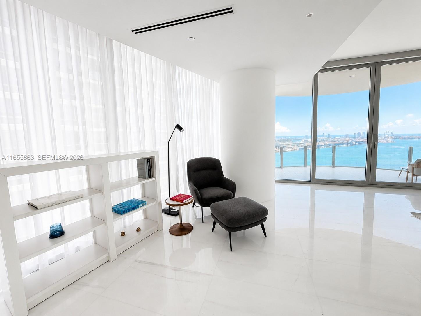A modern living area features a dark armchair, ottoman, bookshelf, and floor-to-ceiling windows overlooking water — 300 Biscayne Blvd Way # 3201, Miami FL 33131, MIAMI, FL