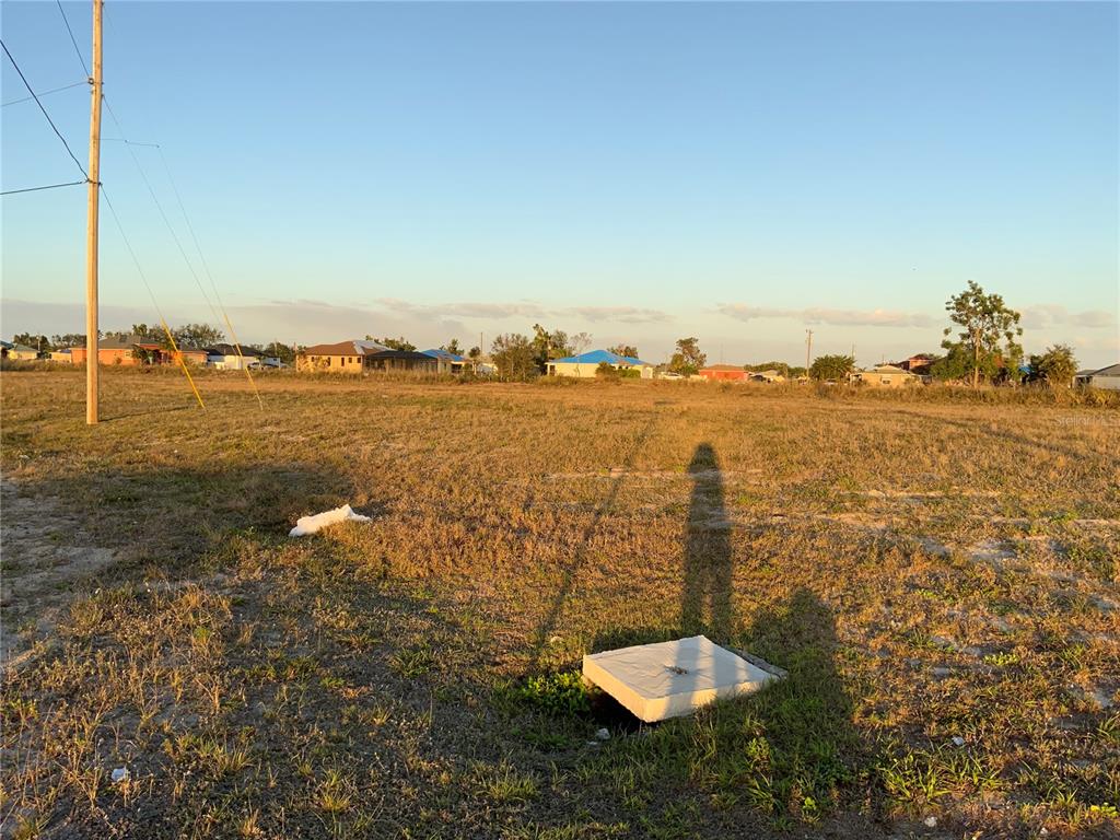 A vacant, grassy lot features a utility cover in the foreground and distant residential houses — 1830 NE 14TH STREET, CAPE CORAL, FL