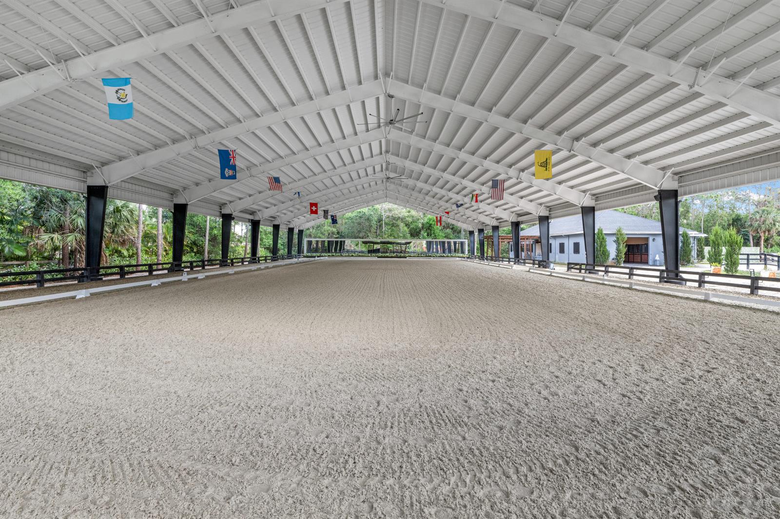 An indoor equestrian riding arena features a sand surface, white metal roof, and hanging flags — 15161 Collecting Canal Road, FL 33470, FL