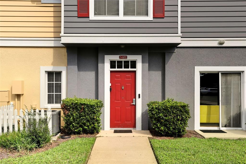 A red front door with a house number sign is centered between two green bushes — 2606 LODI CIRCLE UNIT 102, KISSIMMEE, FL