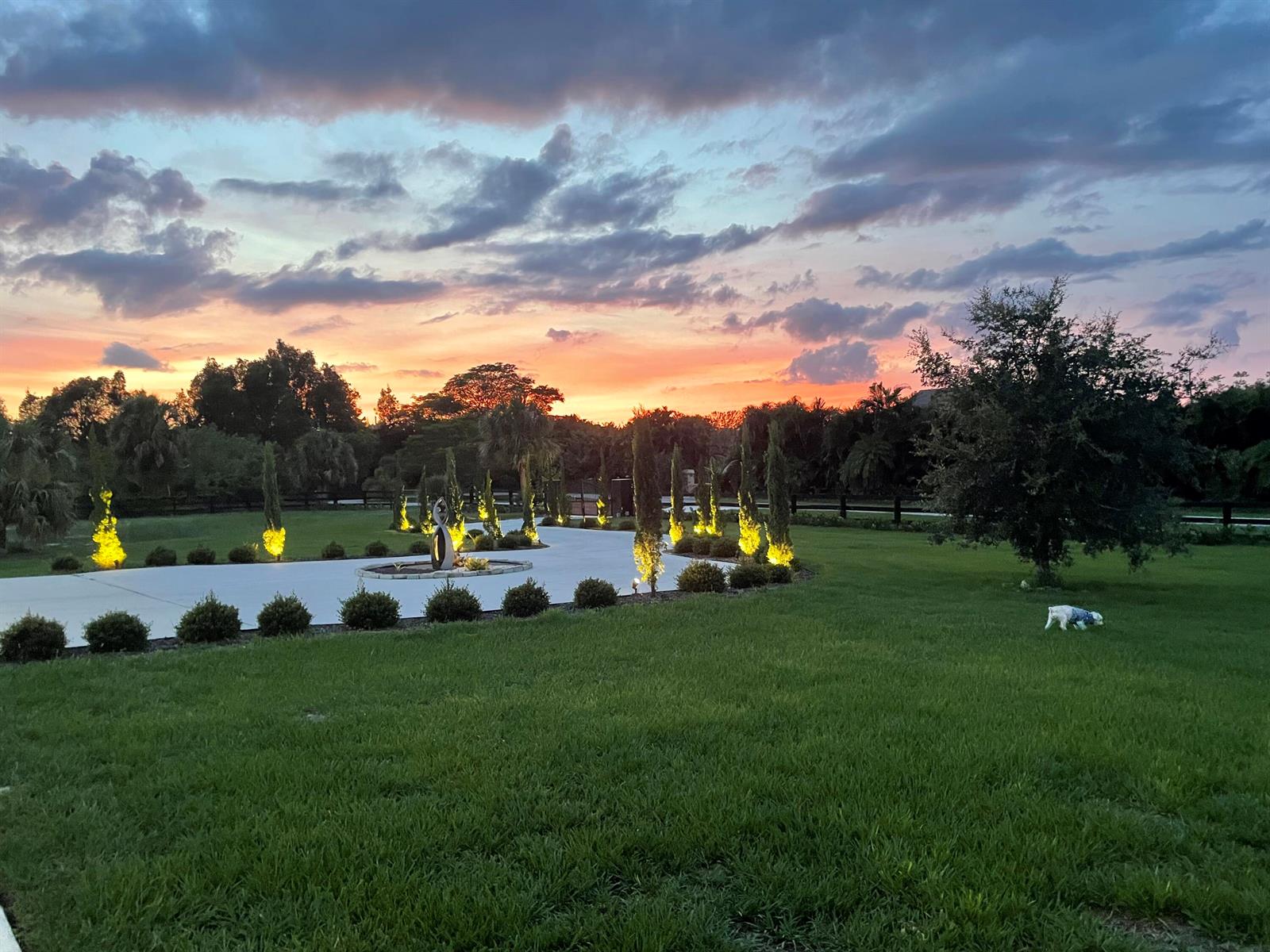 A paved driveway with illuminated landscaping leads toward a sunset sky over a grassy lawn — 19008 Marsh Hawk Lane, Loxahatchee FL 33470, LOXAHATCHEE, FL