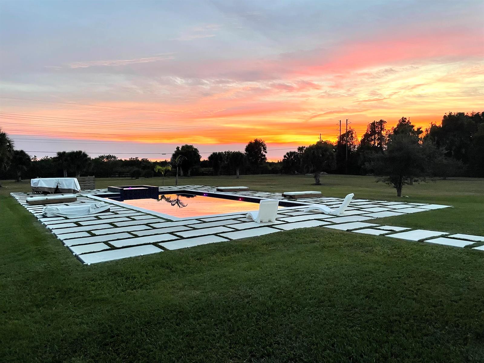 A rectangular swimming pool with a stone patio sits in a grassy yard at sunset — 19008 Marsh Hawk Lane, Loxahatchee FL 33470, LOXAHATCHEE, FL