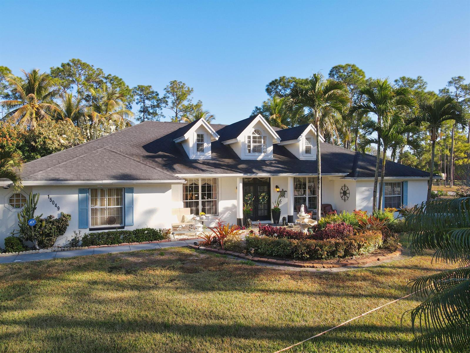 A single-story white house with a dark roof, dormer windows, and a landscaped front yard — 15049 85th Road N, Loxahatchee FL 33470, LOXAHATCHEE, FL