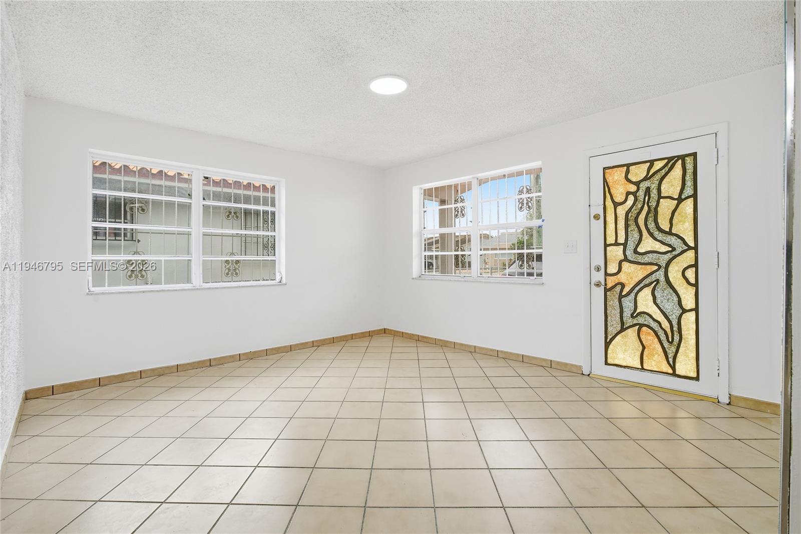 A room with white walls, beige tile flooring, two windows with security bars, and a decorative door — 836 W 37th St, Hialeah FL 33012, HIALEAH, FL