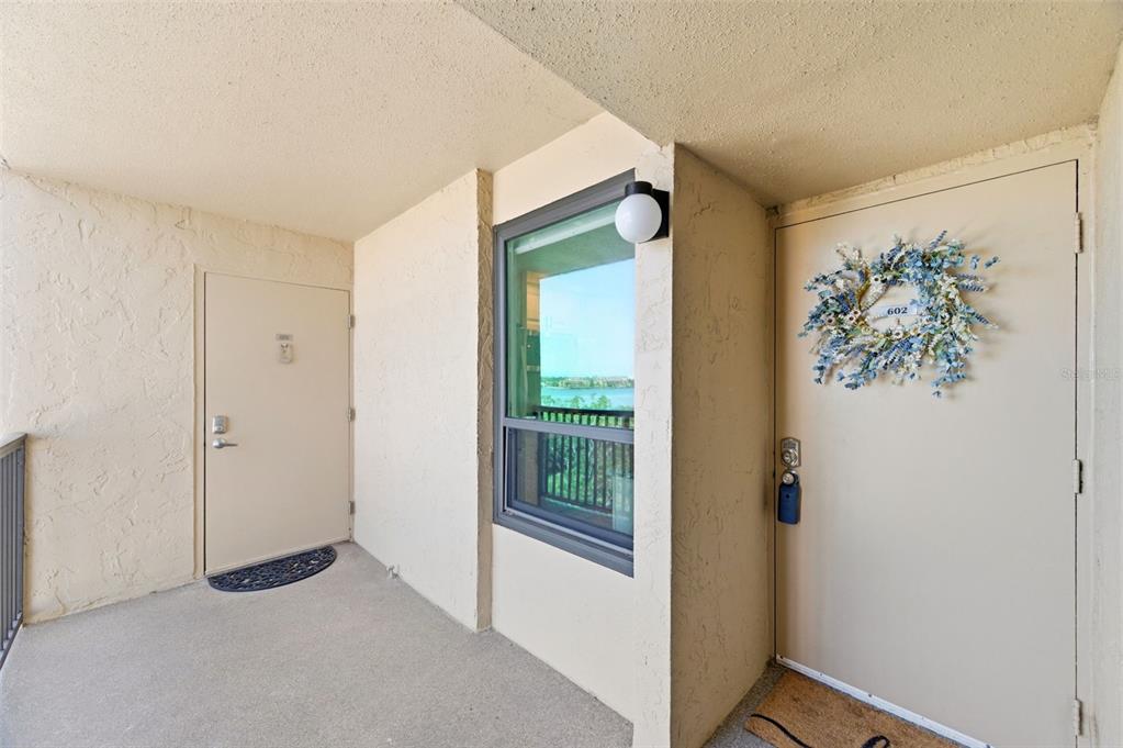 An outdoor apartment hallway features two beige doors, a window, and a wall-mounted light fixture — 2616 COVE CAY DRIVE UNIT 602, CLEARWATER, FL