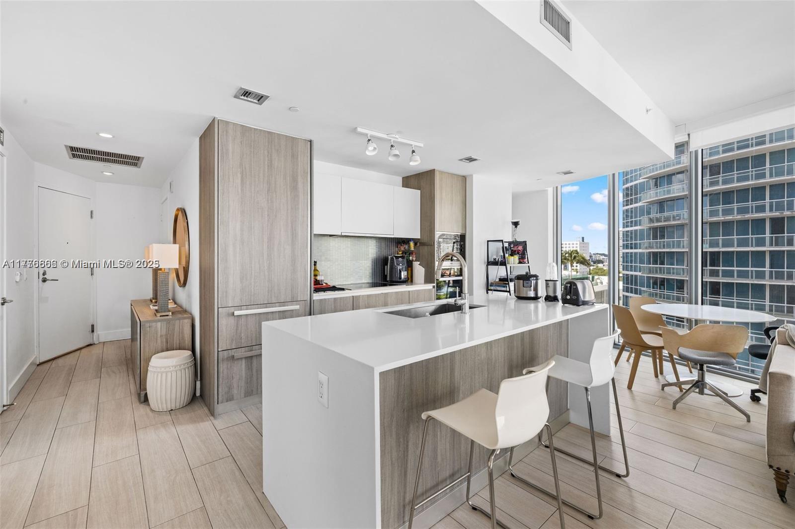 A modern kitchen features a white island, wood-toned cabinetry, and floor-to-ceiling windows with city views — 2900 NE 7th Ave # 901, Miami FL 33137, MIAMI, FL