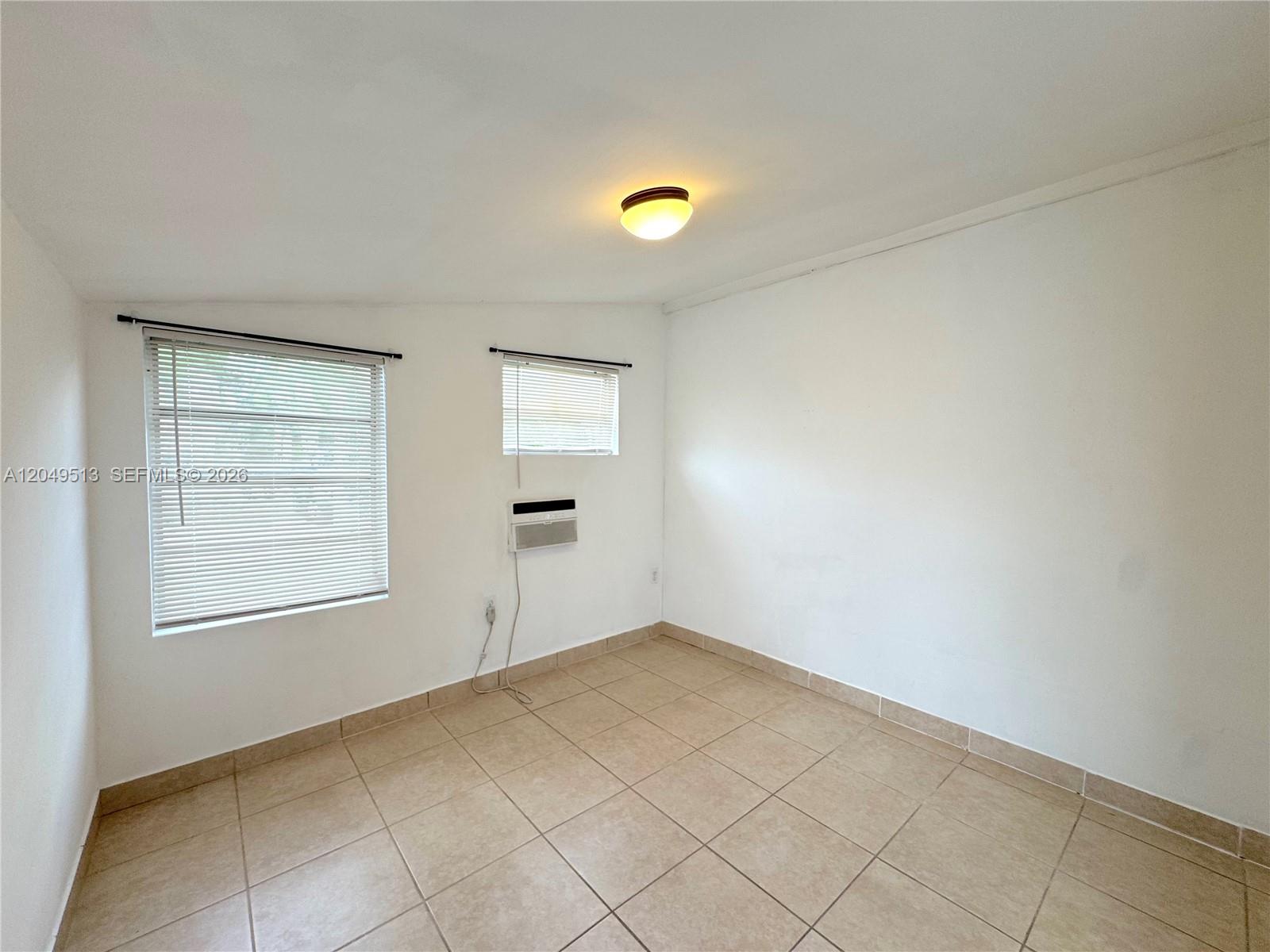 An empty room features white walls, beige tile flooring, two windows with blinds, and an air conditioner — 2461 NW 5th St # C, Miami FL 33125, MIAMI, FL