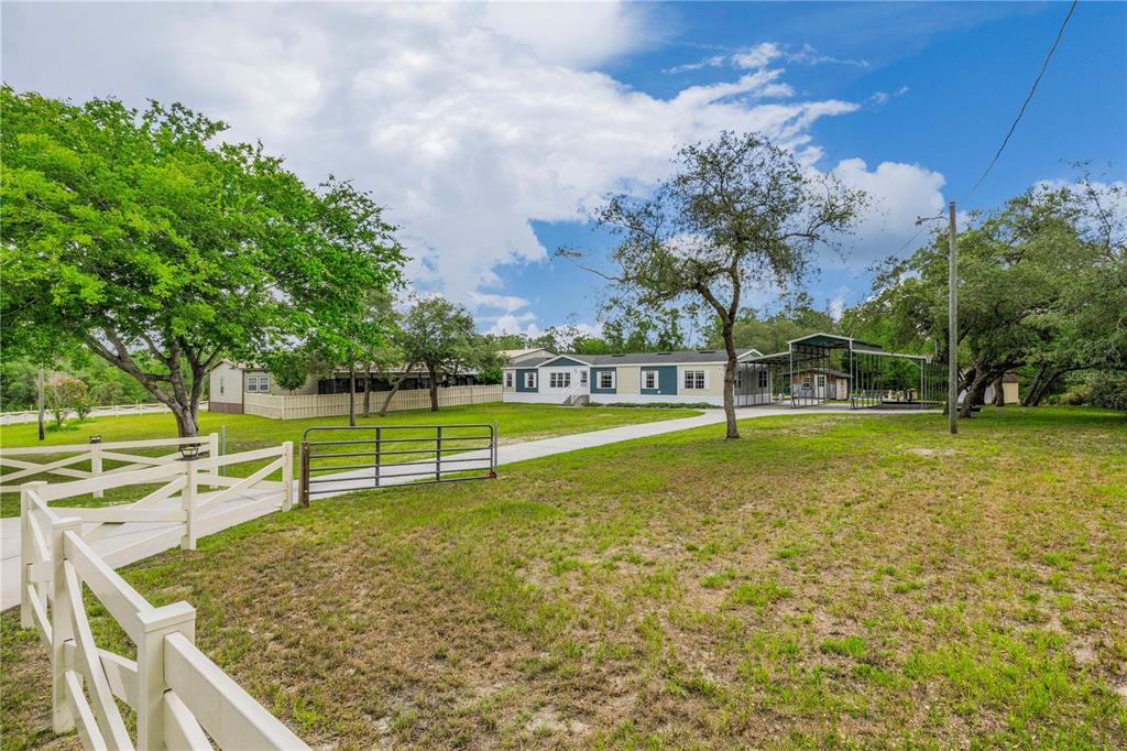 A single-story manufactured home with a metal carport sits on a grassy lot with fencing — 6311 NODOC ROAD, SPRING HILL, FL