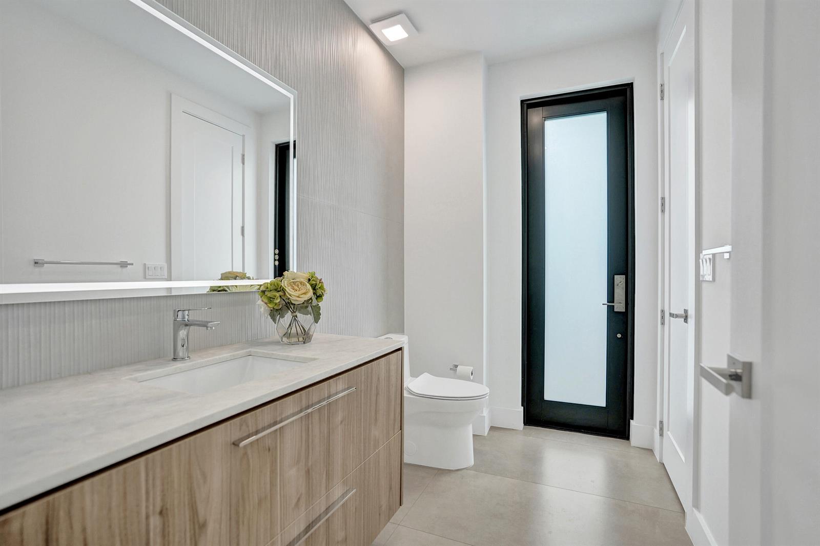 A modern bathroom features a wood vanity, white countertop, large mirror, and a frosted glass door — 235 NE 11th Street, Delray Beach FL 33444, DELRAY BEACH, FL