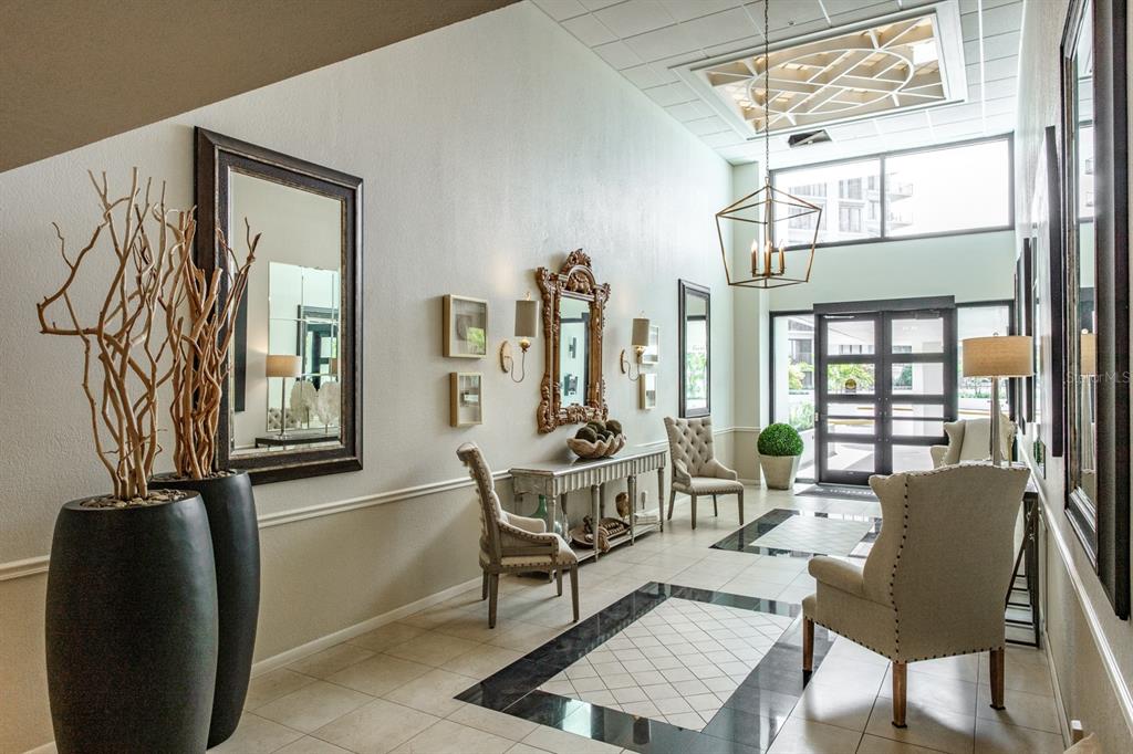 A bright, high-ceilinged lobby features tiled floors, decorative mirrors, accent chairs, and a geometric chandelier — 8 BELLEVIEW BOULEVARD UNIT 108, BELLEAIR, FL