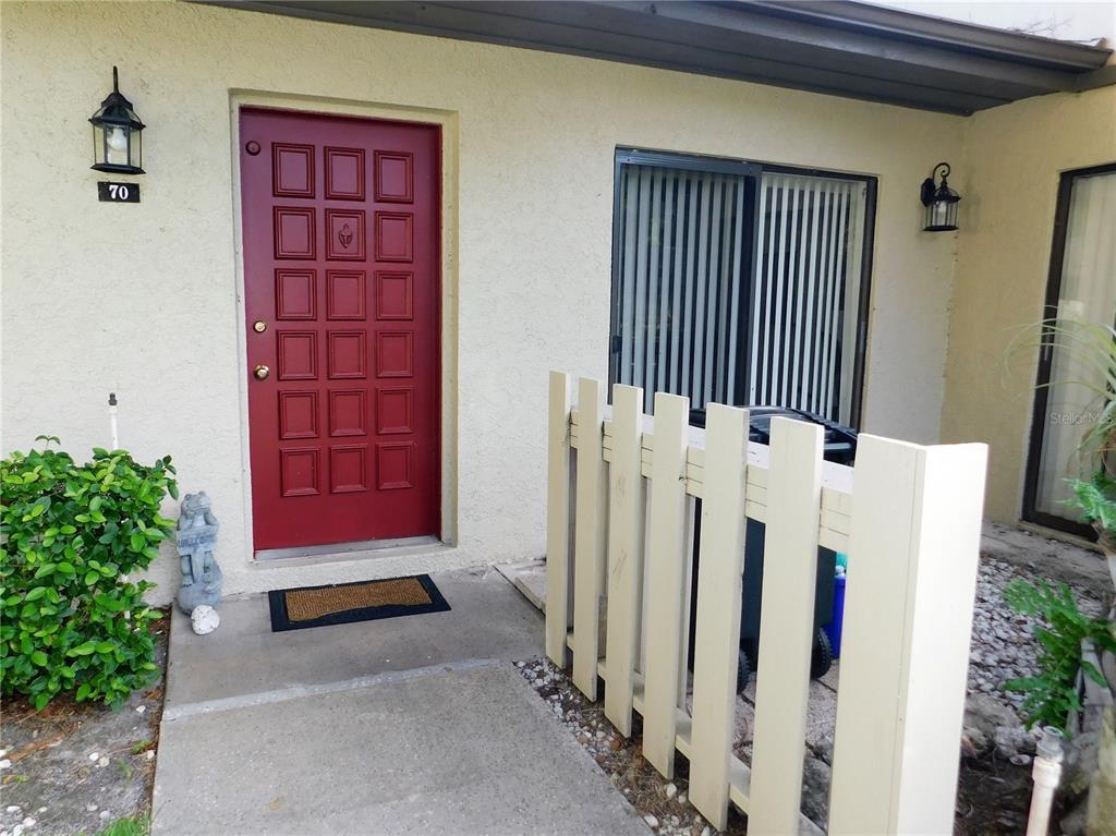 A red front door with a small fence and a trash bin on a concrete patio — 70 JOANNE PLACE UNIT 70, OLDSMAR, FL