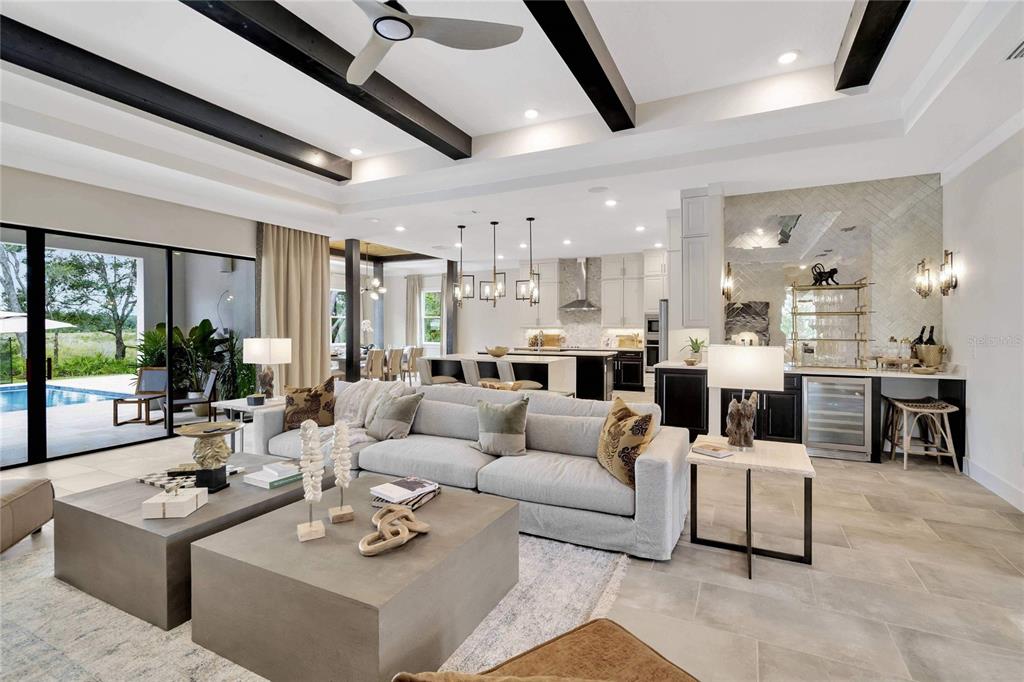 An open-concept living room features a large gray sofa, coffered ceiling, and a kitchen area — 12227 WHITE BLOSSOM COURT, PARRISH, FL