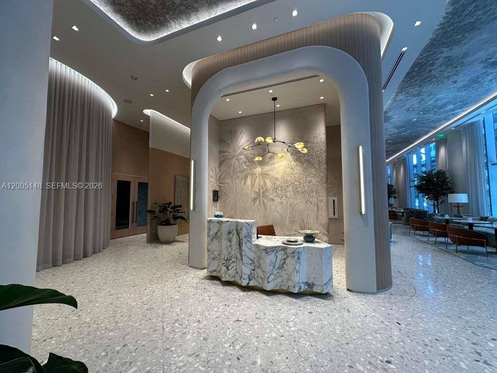 A modern lobby features a marble reception desk, curved architectural elements, and a decorative chandelier — 77 SE 5th Street # 1815, Miami FL 33131, MIAMI, FL