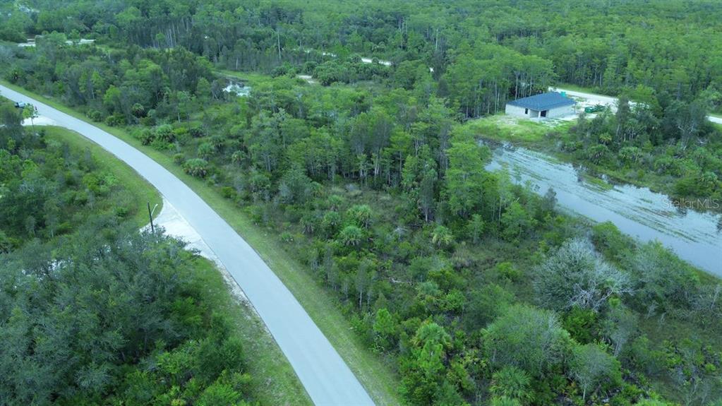 An aerial view shows a paved road curving past a wooded lot with a canal — 486 WESTCHESTER DRIVE, LEHIGH ACRES, FL