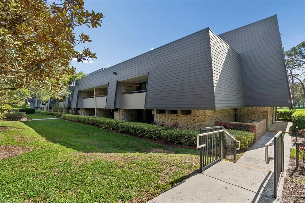 A multi-story building features gray horizontal siding, brick lower walls, and a concrete walkway — 36750 US HIGHWAY 19 UNIT 21214, PALM HARBOR, FL