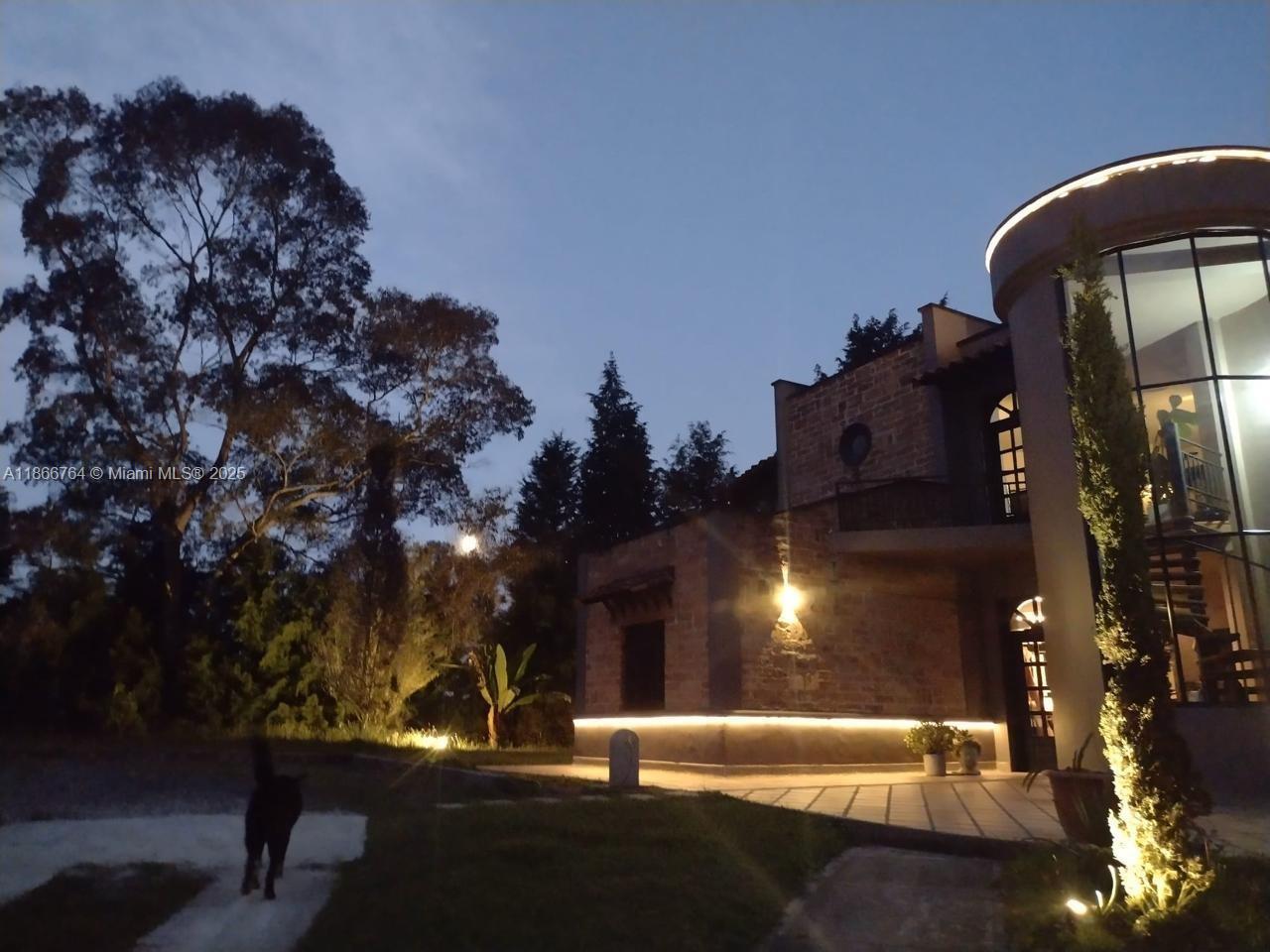 A stone house with a curved glass tower is illuminated at dusk near a large tree — Lote 4 Vereda La Honda, At 054, At