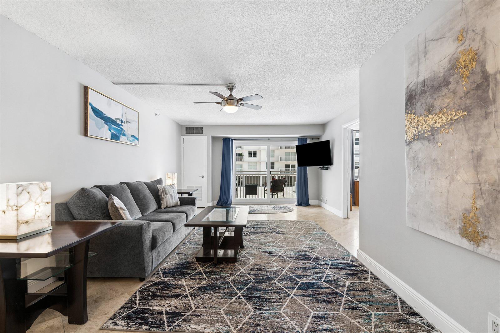 A living room features a gray sofa, patterned area rug, ceiling fan, and balcony access — 4511 S Ocean Boulevard # 106, Highland Beach FL 33487, HIGHLAND BEACH, FL