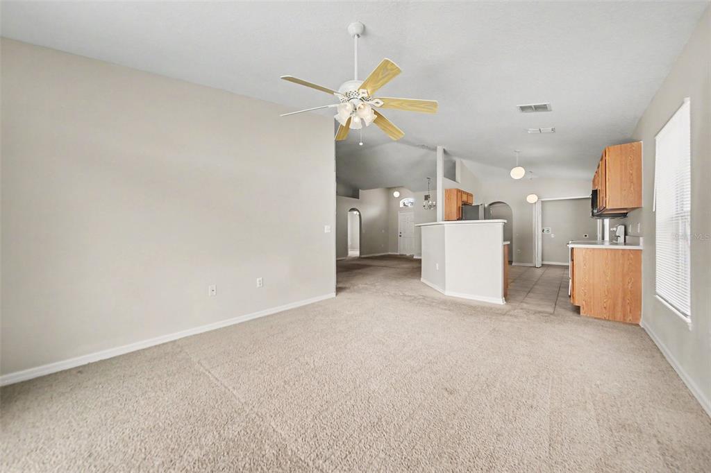 A living room with beige carpet and a ceiling fan opens to a kitchen area — 2122 AITKIN LOOP, LEESBURG, FL