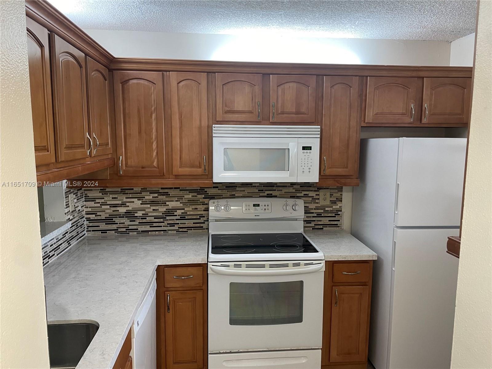A kitchen features wood cabinets, white appliances, a mosaic tile backsplash, and light countertops — 3252 Foxcroft Rd # 206, Miramar FL 33025, MIRAMAR, FL