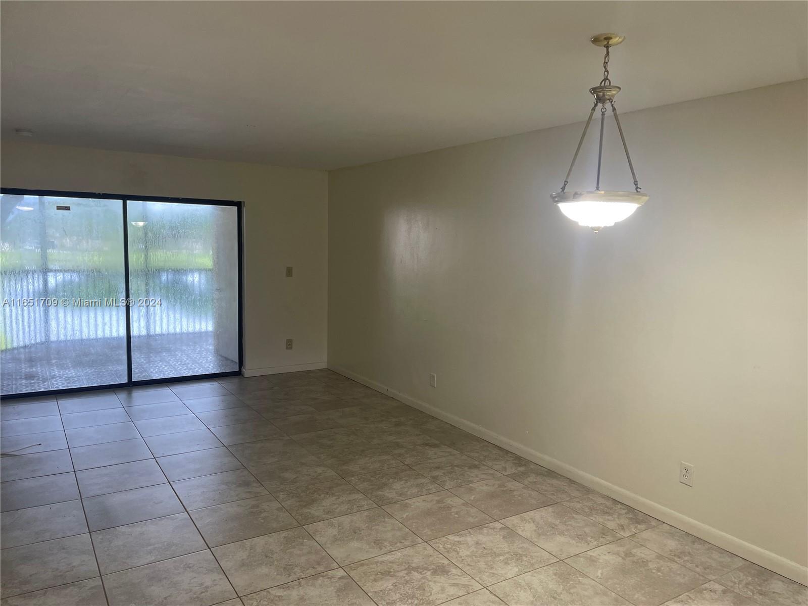 An empty room features light-colored tile flooring, a hanging light fixture, and sliding glass doors — 3252 Foxcroft Rd # 206, Miramar FL 33025, MIRAMAR, FL