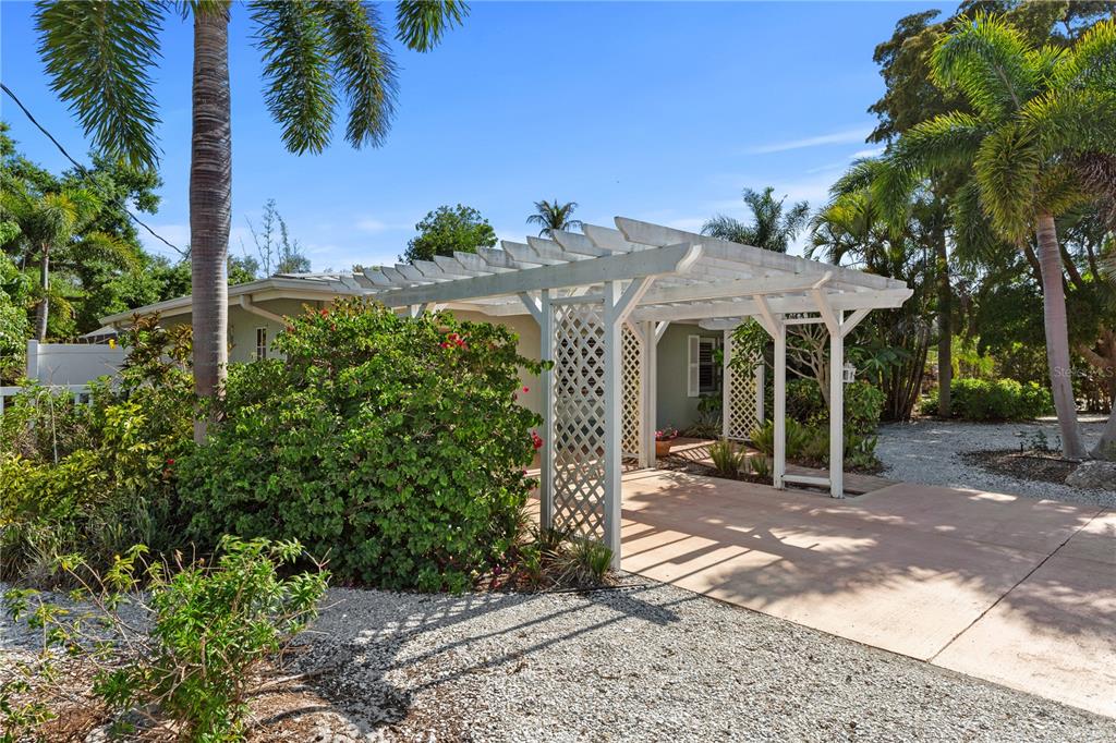 A white wooden pergola stands over a concrete driveway next to a house with landscaping — 7860 HOLIDAY DRIVE, SARASOTA, FL