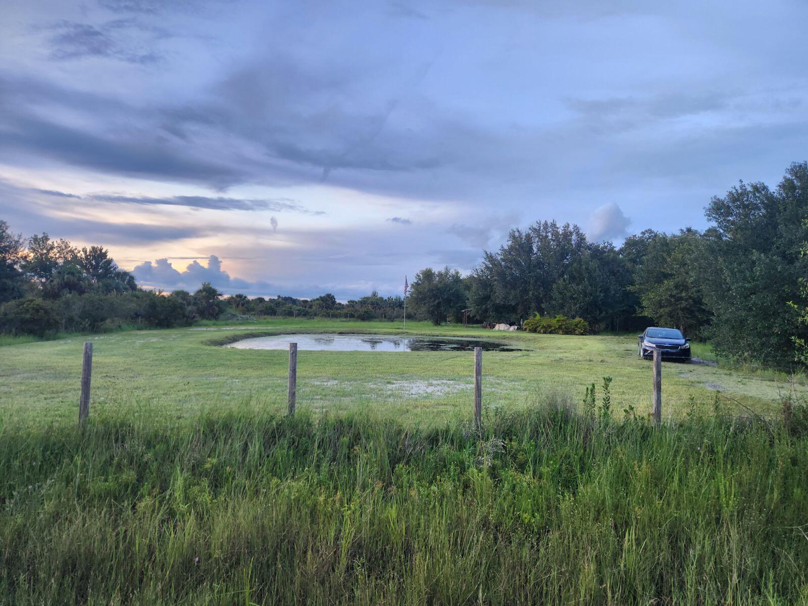 A grassy field features a small pond, a parked car, and a wooden fence — 20641 NW 302nd Street, Okeechobee FL 34972, OKEECHOBEE, FL