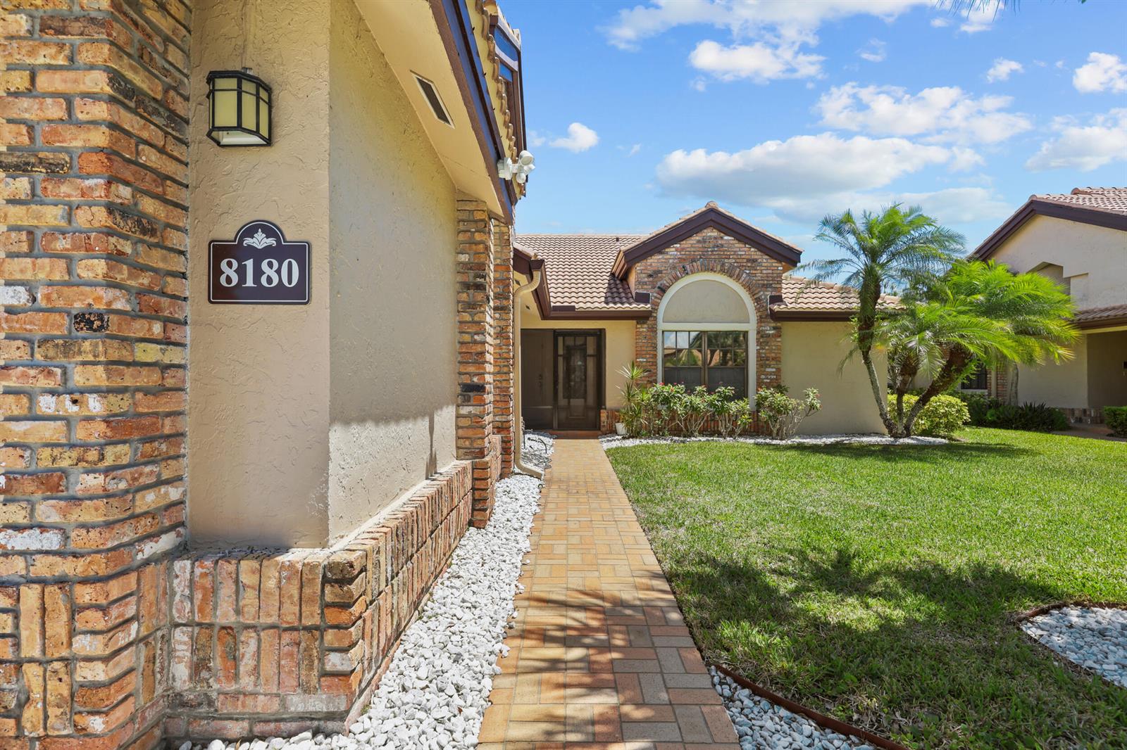 A tan stucco house exterior features a brick-accented wall, a paved walkway, and landscaping — 8180 Bellwort Place, Boynton Beach FL 33472, BOYNTON BEACH, FL