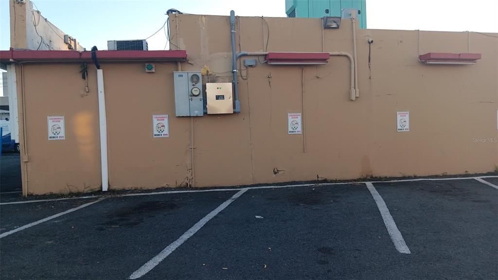A tan exterior wall of a commercial building features electrical equipment and parking space markings — PR-167 KM 16.1 BUENA VISTA WARD - FALCON SECTOR, BAYAMON, PR