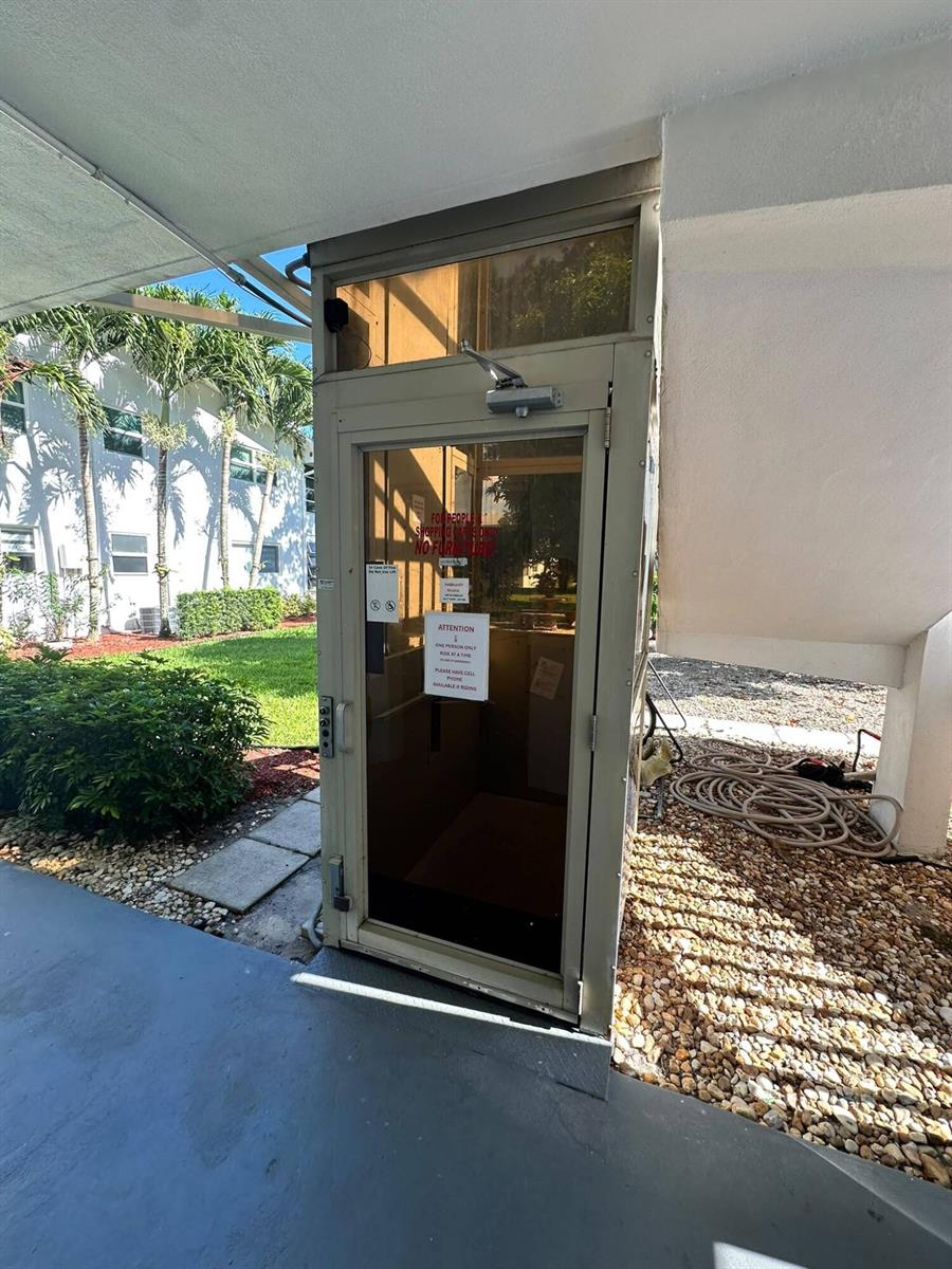 An outdoor elevator enclosure with a glass door stands on a concrete walkway — 171 Markham H # 171, Deerfield Beach FL 33442, DEERFIELD BEACH, FL