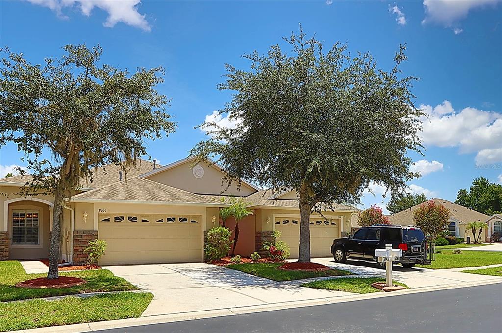 A beige suburban house with two garage doors and mature trees in the front yard — 31007 PROUT COURT, WESLEY CHAPEL, FL