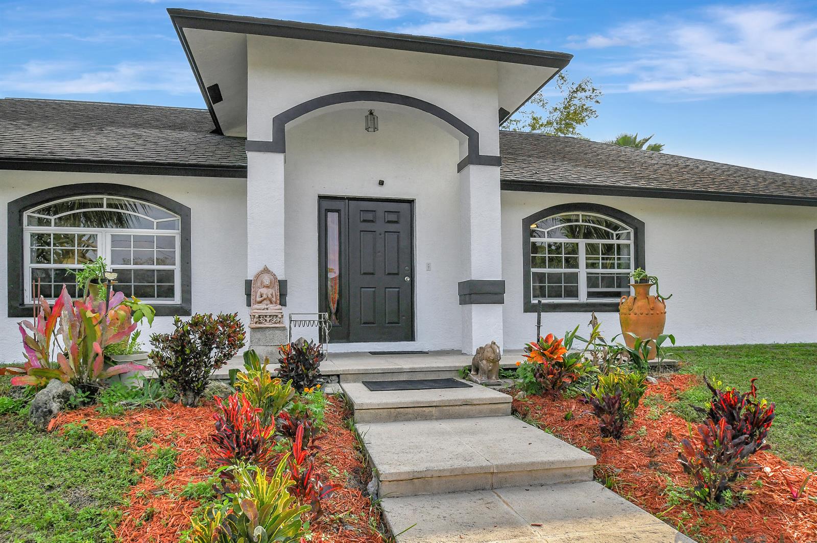 The exterior of a white house features a dark arched entryway, windows, and landscaped gardens — 3446 Cabbage Palm Way, Loxahatchee FL 33470, LOXAHATCHEE, FL