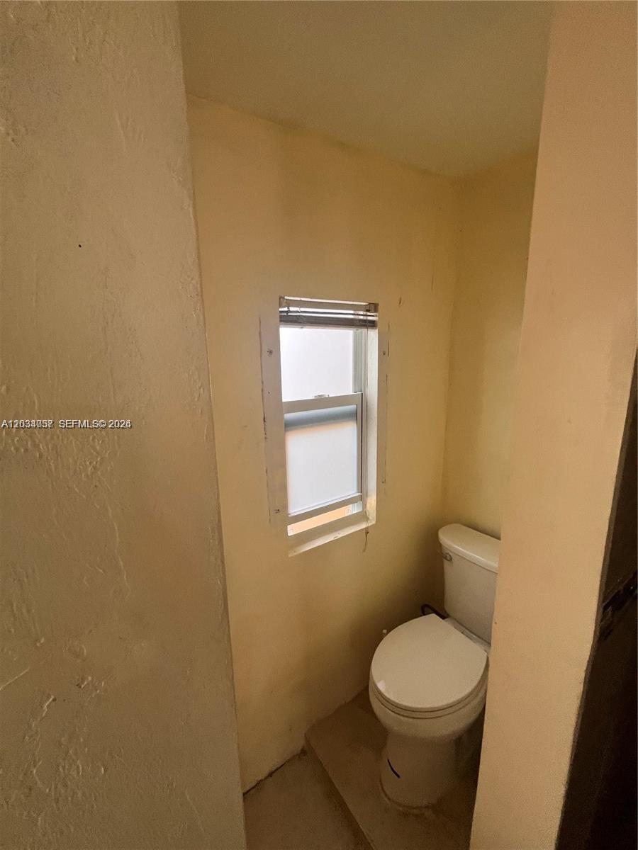 A small bathroom features a white toilet situated beneath a single window with blinds — 13215 NW 19th Ave # 1, Miami FL 33167, MIAMI, FL