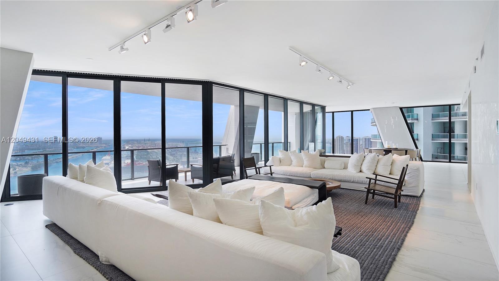 A modern living room features white sofas, track lighting, and floor-to-ceiling windows overlooking the ocean — 1000 Biscayne Blvd # 4401, Miami FL 33132, MIAMI, FL