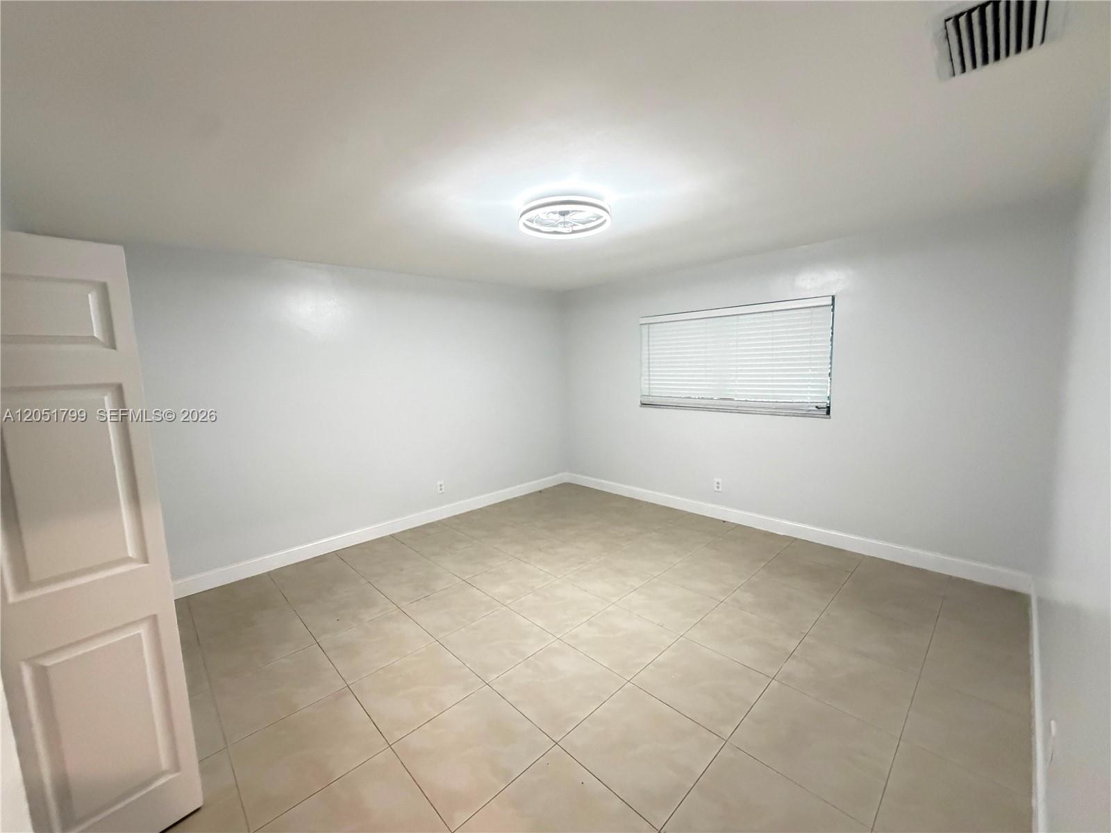 An empty room features light-colored walls, beige tile flooring, a ceiling light, and a window — HOLLYWOOD, FL