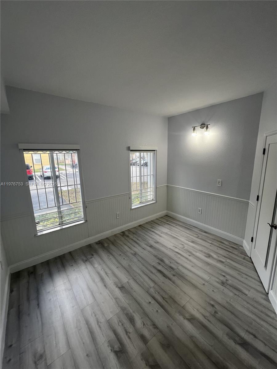 An empty room features gray wood-look flooring, white wainscoting, two windows with security bars, and a wall-mounted light — 9000 W Flagler St # 1, Miami FL 33174, MIAMI, FL