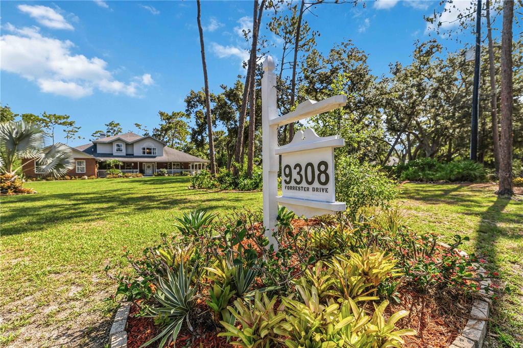 A white sign displaying the number 9308 stands in a landscaped yard before a house — 9308 65TH AVENUE E, BRADENTON, FL
