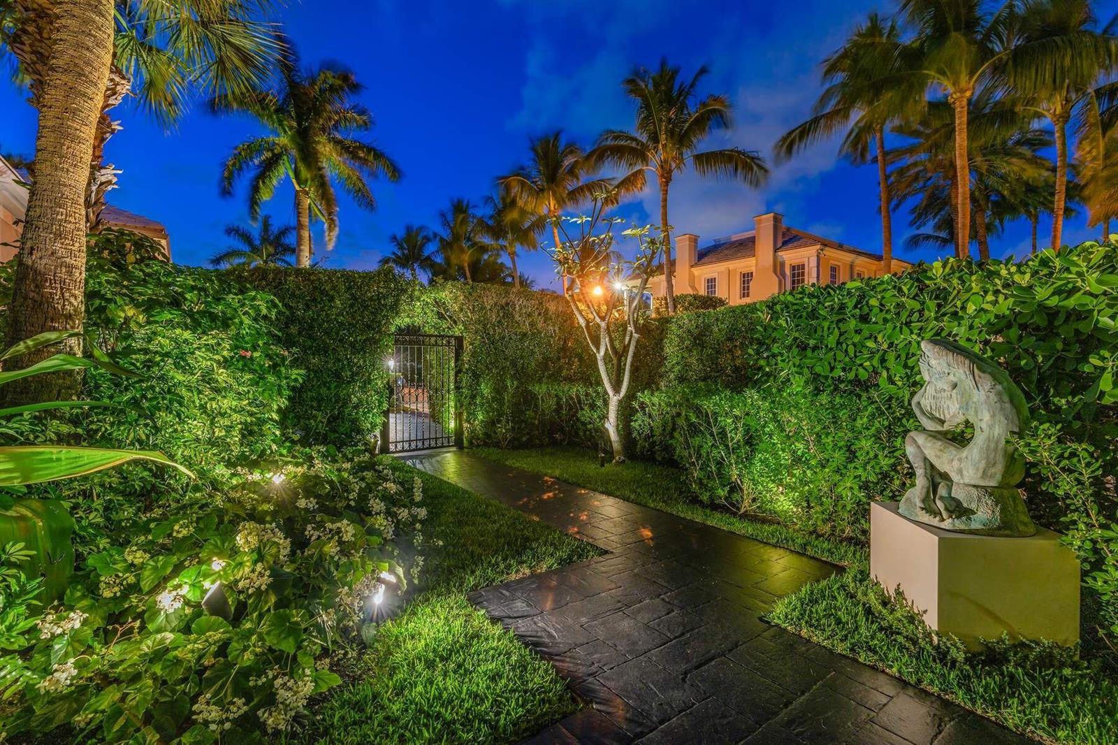A stone walkway leads through a lush, illuminated garden toward a gated entrance at night — 710 N COUNTY Road, Palm Beach FL 33480, PALM BEACH, FL