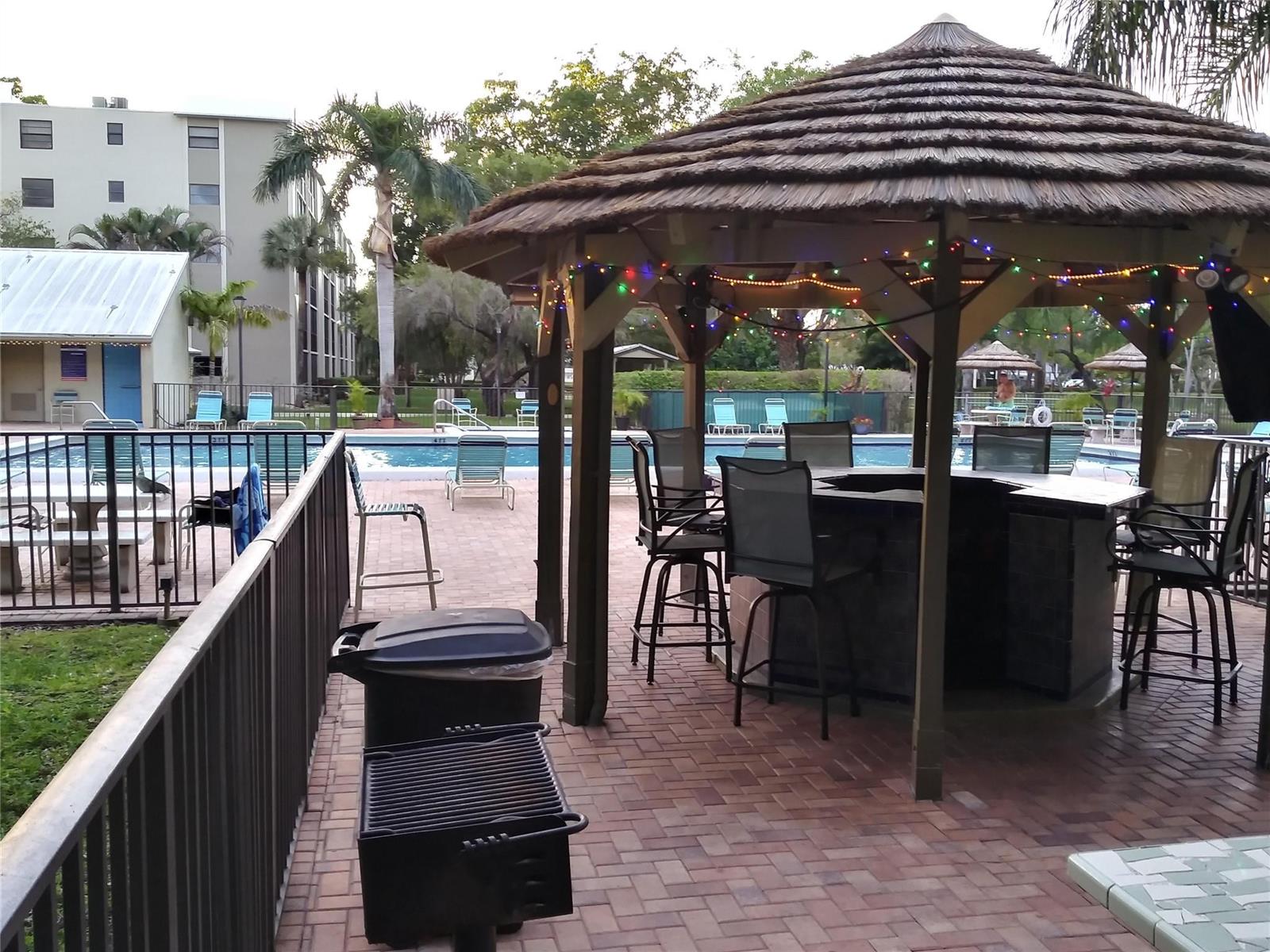 A thatched-roof tiki bar with stools sits on a brick patio beside a swimming pool — 2112 S Cypress Bend Drive # 207, Pompano Beach FL 33069, POMPANO BEACH, FL
