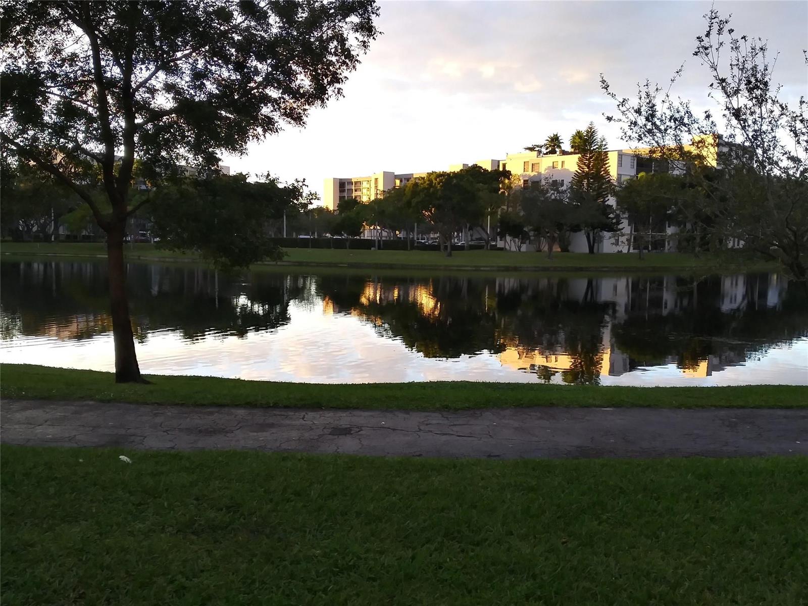 A paved path runs along a grassy bank overlooking a pond with apartment buildings beyond — 2112 S Cypress Bend Drive # 207, Pompano Beach FL 33069, POMPANO BEACH, FL