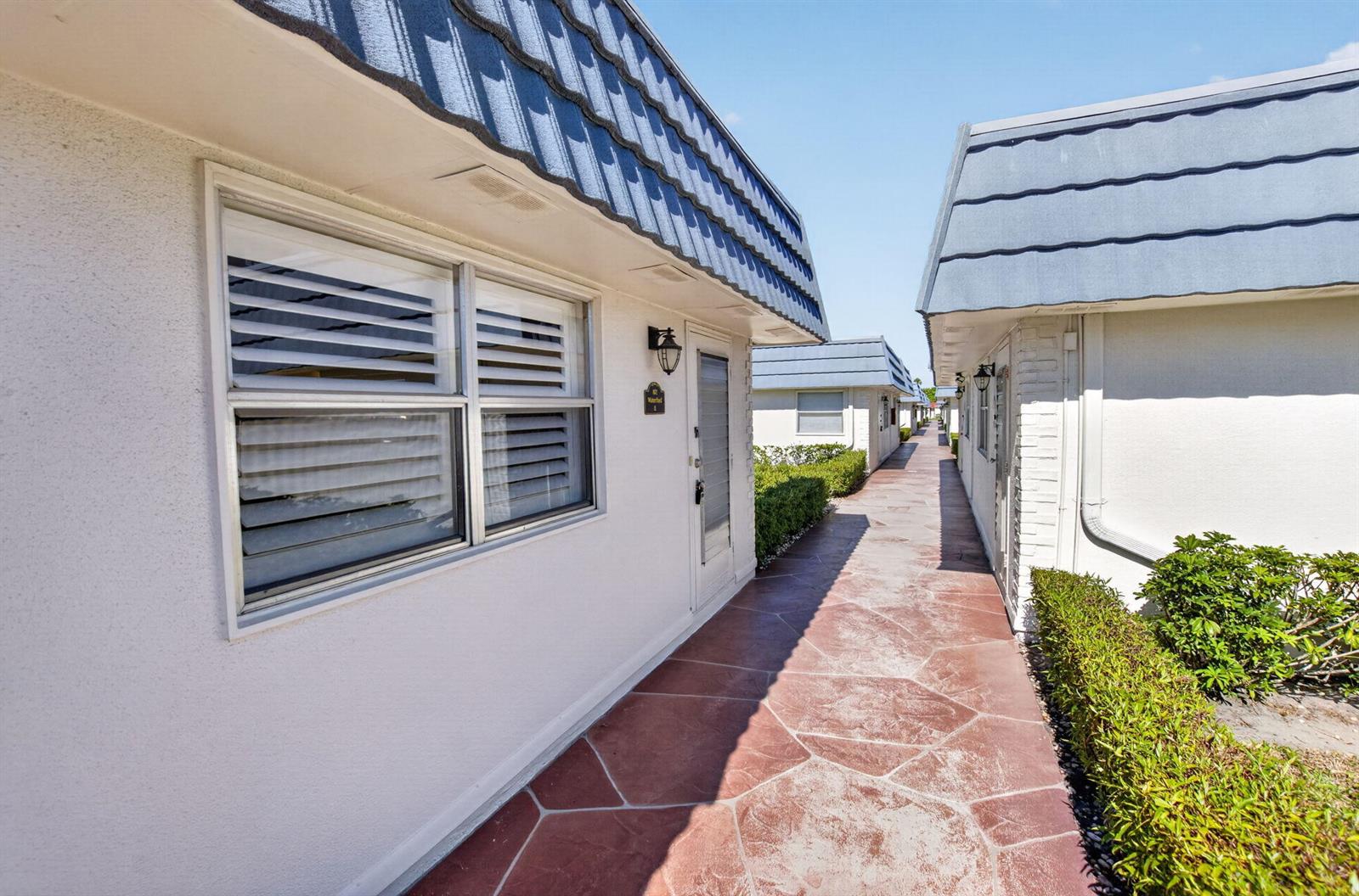 A narrow outdoor walkway separates two rows of single-story buildings with blue mansard roofs — 102 E Waterford E # 102, Delray Beach FL 33446, DELRAY BEACH, FL