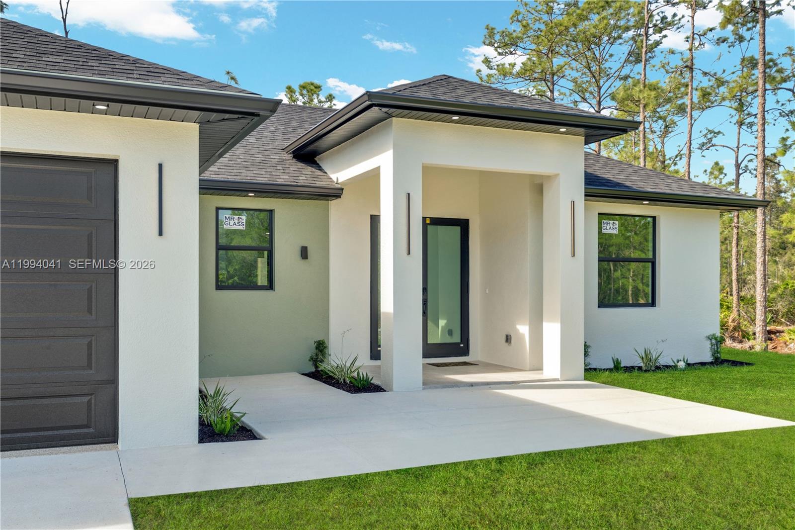 The exterior of a modern house features a dark garage door, white walls, and landscaping — 2102 E 13th St, Lehigh Acres FL 33972, LEHIGH ACRES, FL