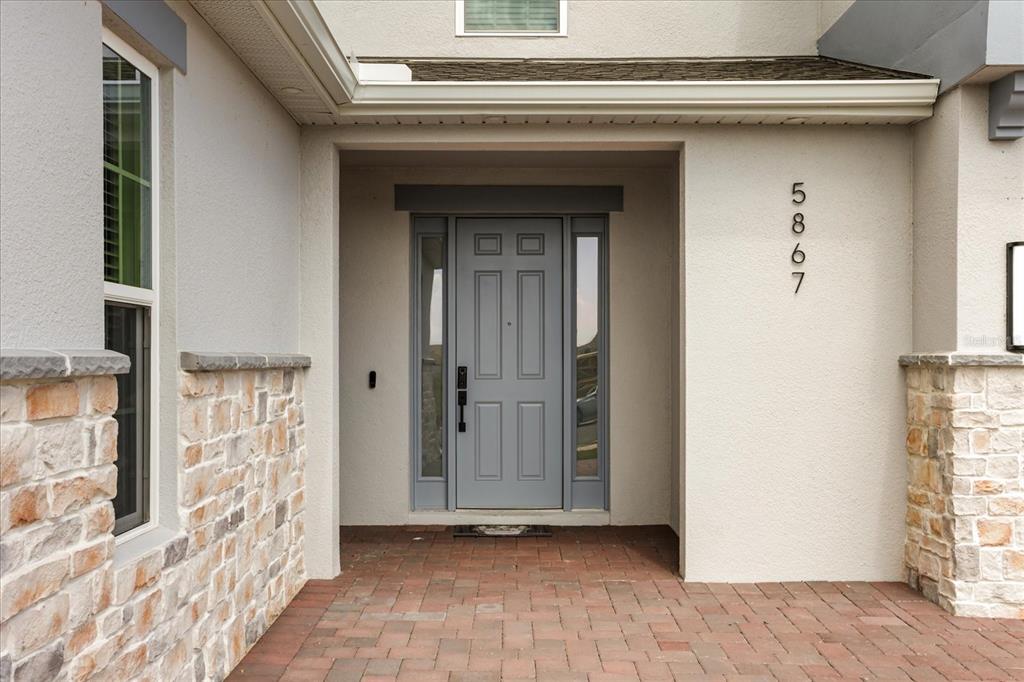 A gray front door with sidelights is framed by stone veneer and a brick walkway — 5867 SIERRA CROWN STREET, MOUNT DORA, FL