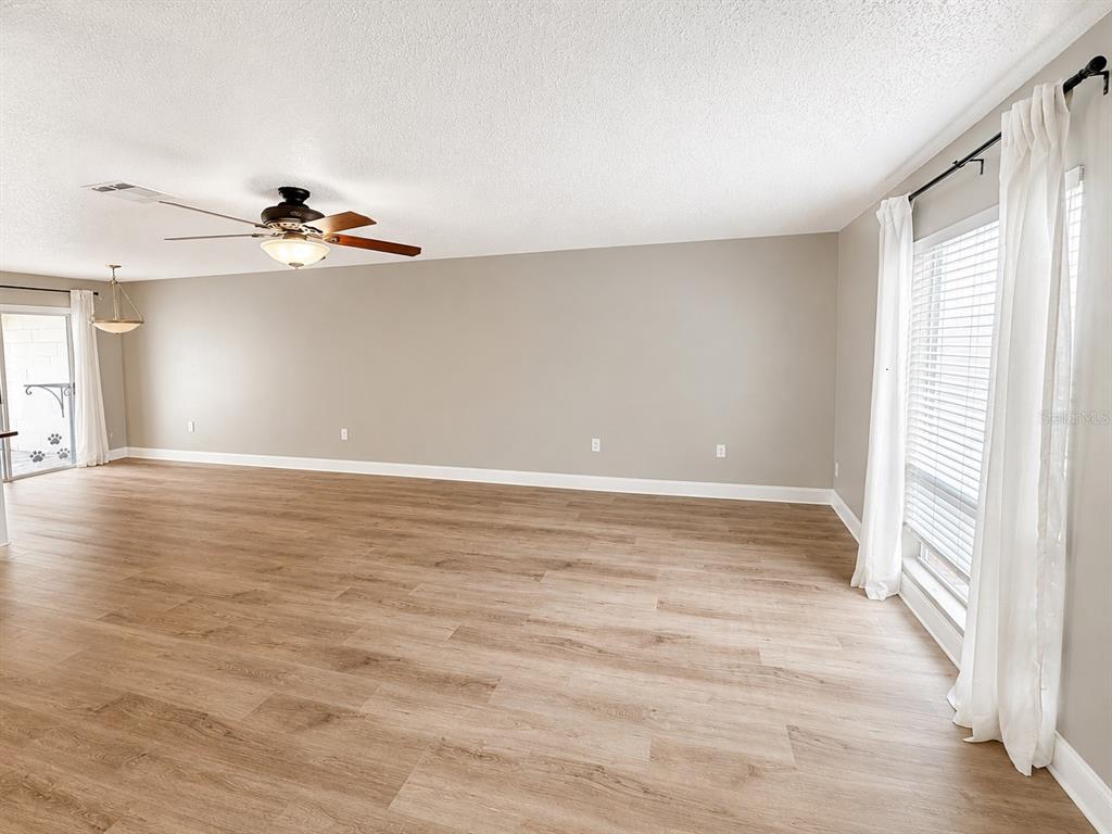 An empty living room features light wood-look flooring, beige walls, a ceiling fan, and large windows — 11905 CYPRESS VISTA, TAMPA, FL