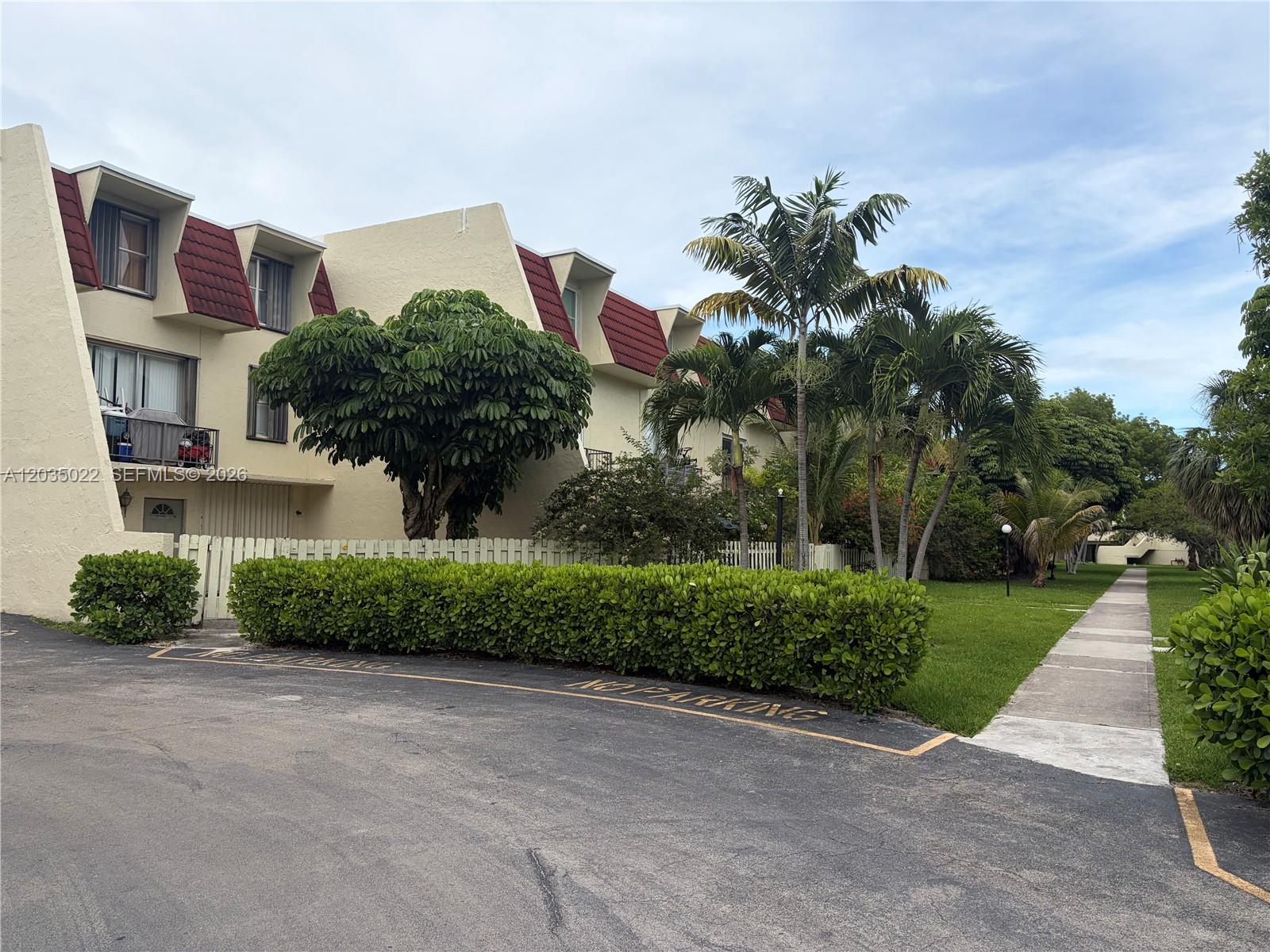 A multi-story apartment building with red mansard roofs sits behind a hedge and palm trees — 1655 NE 115th St # 29B, Miami FL 33181, MIAMI, FL