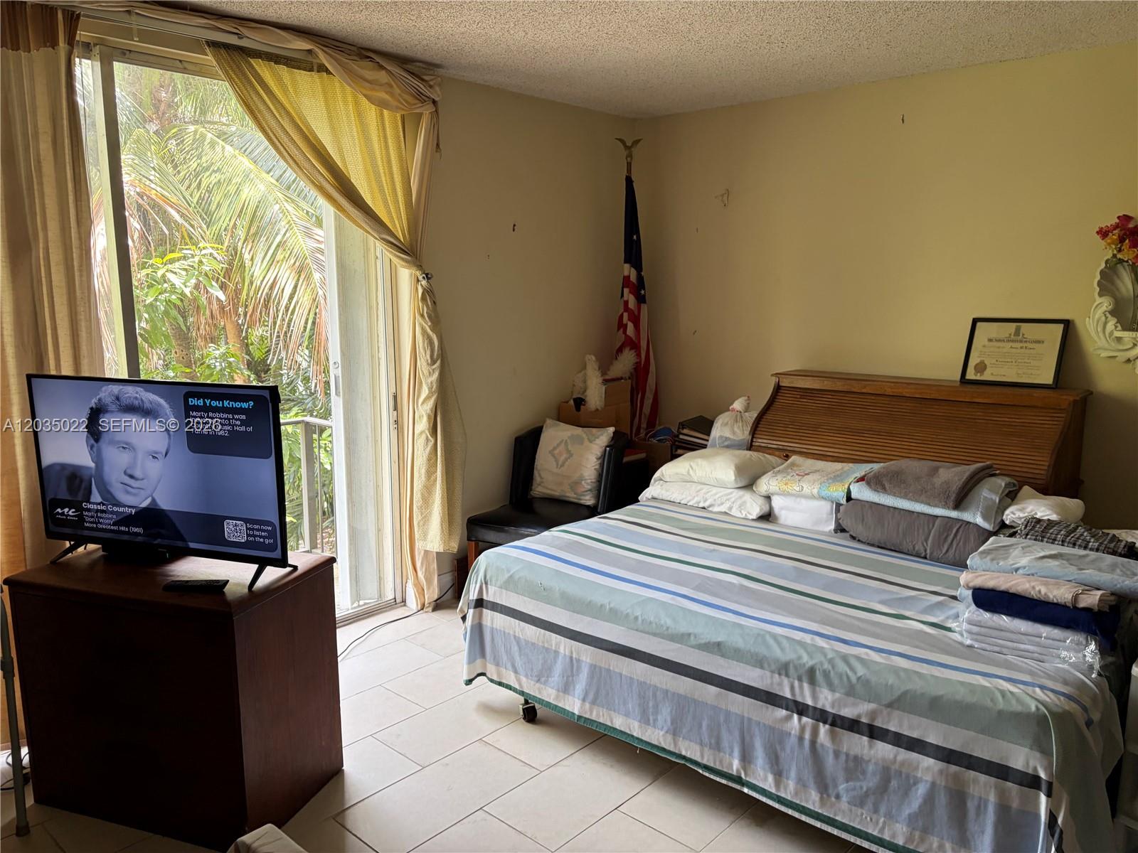 A bedroom features a bed with striped bedding, a television on a stand, and a balcony — 1655 NE 115th St # 29B, Miami FL 33181, MIAMI, FL