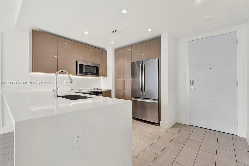 A modern kitchen features white countertops, glossy taupe cabinetry, stainless steel appliances, and tile flooring — 2000 Metropica Way # 2307, Sunrise FL 33323, SUNRISE, FL